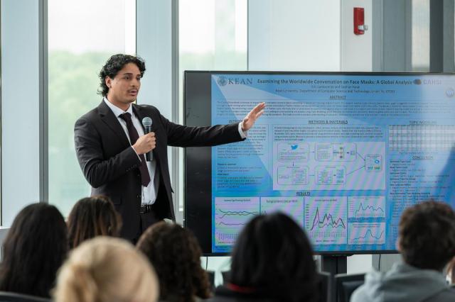 Eric Landaverde, in a jacket and tie, points to a his research on a large screen.