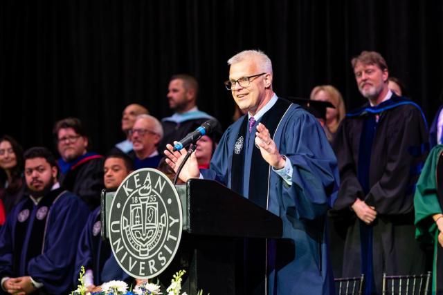 Kean Board Chair Steve Fastook in blue commencement gown speaks at a podium.