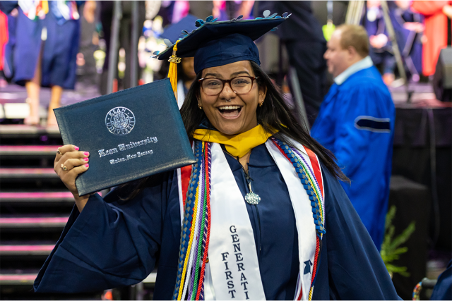 A young woman in a Kean cap and gown holds her diploma cover and smiles. She wears a stole reading, First Generation.