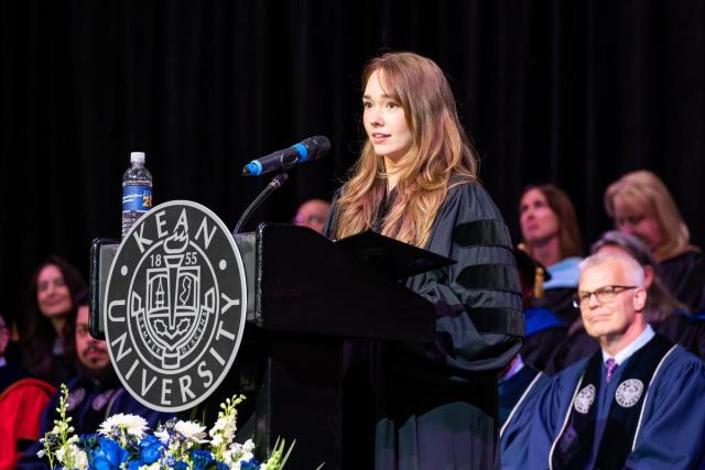 Actor Holly Taylor in a blue commencement gown speaks at a podium.