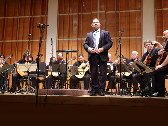 A group of musicians on stage, members of the New Jersey Guitar Orchestra, one man in a suit is standing up and the shot is from audience view