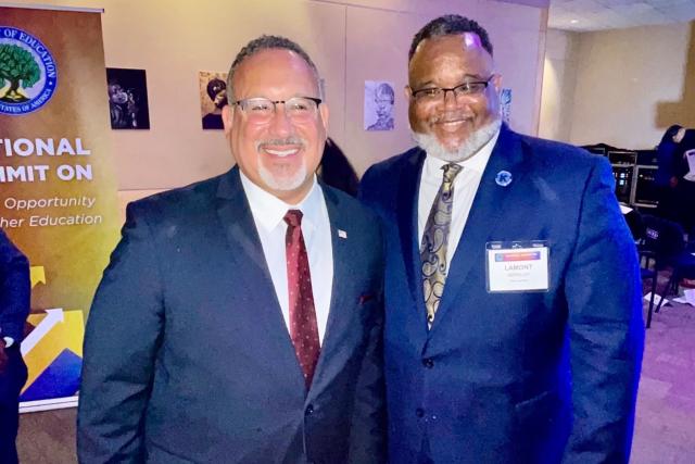 Two mean wearing blue suits; one Hispanic, one Black, stand shoulder to shoulder, smiling.