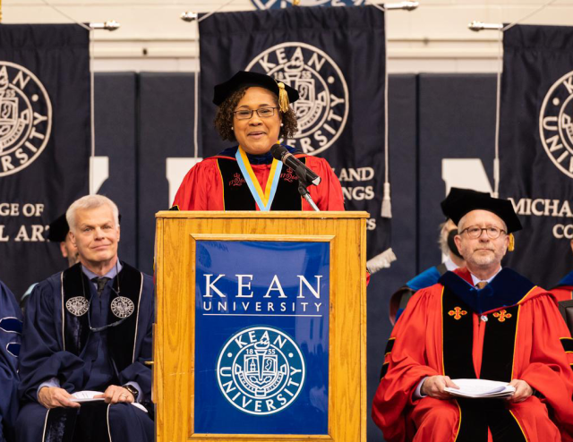 Rajade Berry-James, in a red commencement robe, speaks from a Kean podium at Harwood Arena.