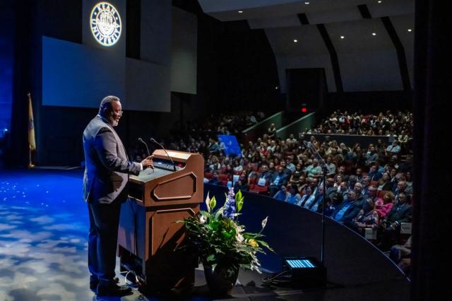 A side view of Kean University Repollet at the Wilkins Theatre podium, with the audience in front of him and the Kean seal projected on a wall.