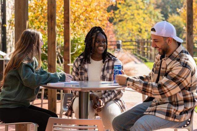 Three students at a round table laugh. A bridge and falll foliage are behind them.