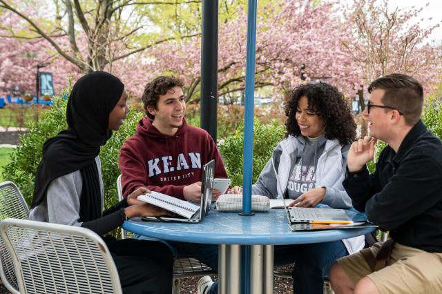 Four students, two women and two men, sit at a table with their laptops, outdoors on Kean's Union campus