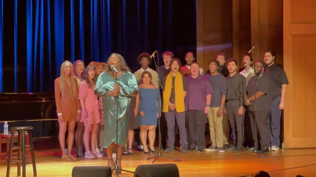 Alex Newell, in a green dress, sings onstage, with Kean students singing behind them.