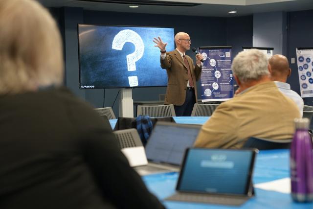 Dr. Henning at the front of a room gestures, while an audience seated at tables listens.