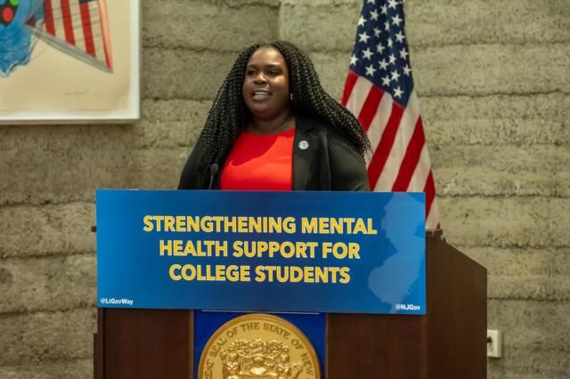 Itunu Balogun, in a red dress and black jacket, at a podium with the sign Strengthening Mental Health Support for College Students
