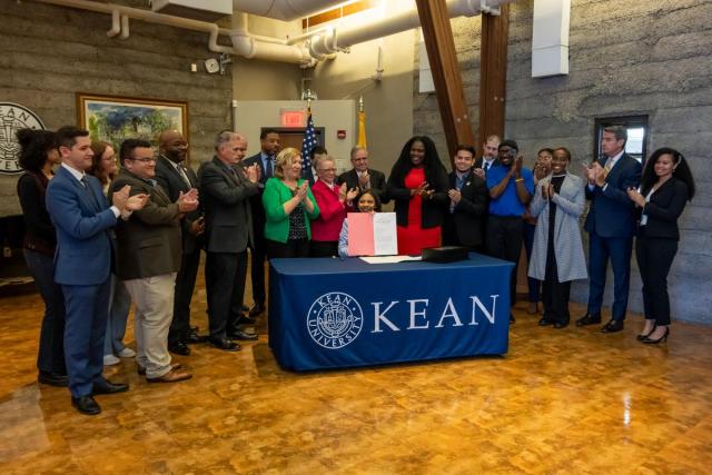 Acting Governor Tahesha Way signs a bill at a table with the Kean seal, as people standing around her applaud.
