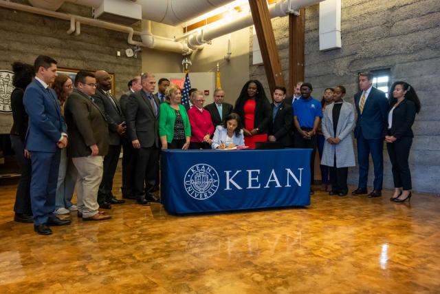 Acting Governor Tahesha Way at a table with the Kean seal signs a bill while a group of students, lawmakers and mental health advocates look on.