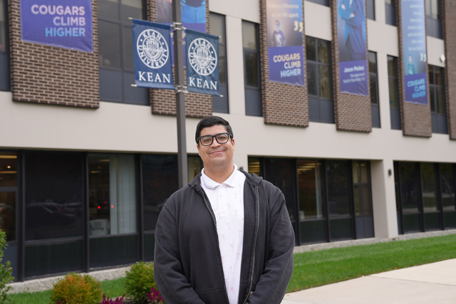 Gabriel Serrano, wearing a black jacket over a white shirt, stands in front of a Kean University building.