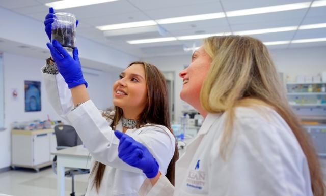 A female student holds a beaker aloft as a female professor looks on.