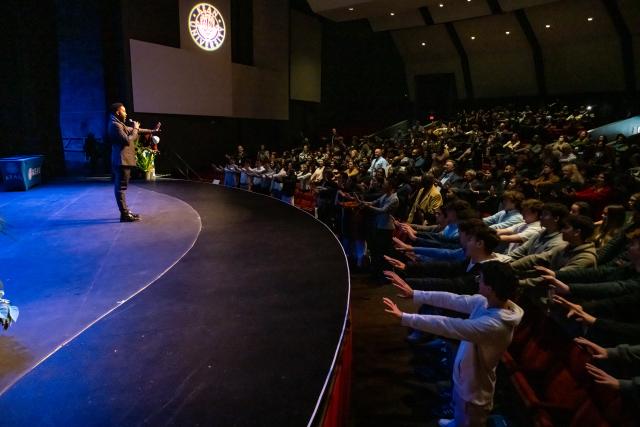 An image from the side of the stage capturing Ndaba Mandela on stage, speaking with his one hand holding the microphone and his other arm out in front of him, and the audience with their arms up in front of them.