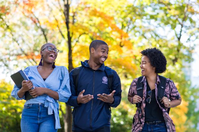 Three students laugh against a backdrop of fall foliage. 