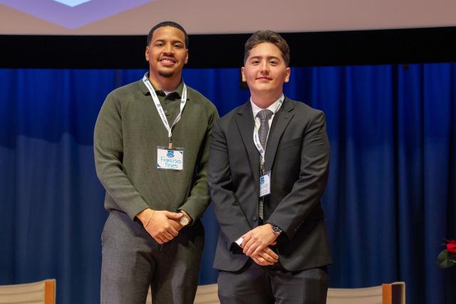 One young black man in a green pullover and a tan young man in a black suit stand on a stage in front of a projector screen