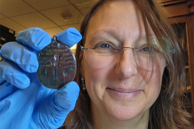 Smiling white female with brown hair, glasses, holding up an amulet in a blue-gloved hand.