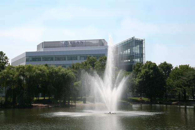 An active fountain in the middle of a small lake lined with trees; multi-level building with windows in the background.