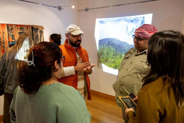 a man in a tan hat and orange vest talks to a group of three women in the middle of an art gallery