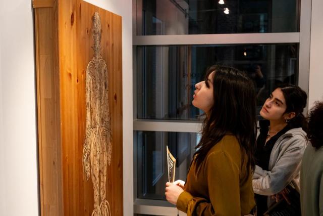 Two girls with dark hair looking at a piece of artwork that is brown and next to a window