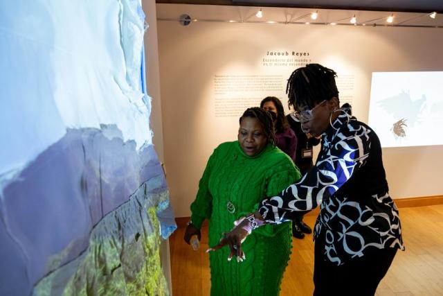 Two women looking into a piece of artwork, one wearing a green dress, one wearing a blue pattern blouse