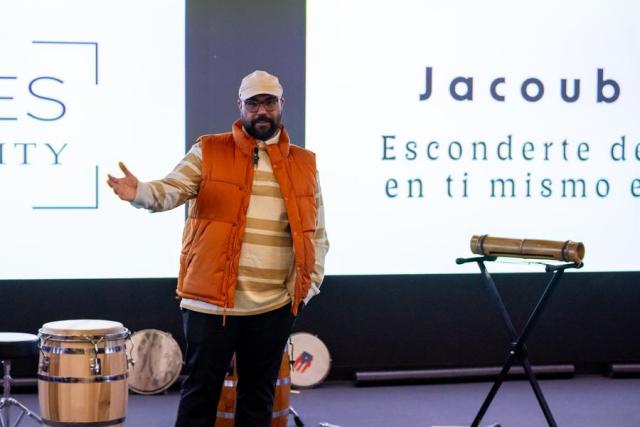 Man wearing glasses, and a hat, with a beard, orange jacket, stripe long sleeve shirt, and jeans with arms extended