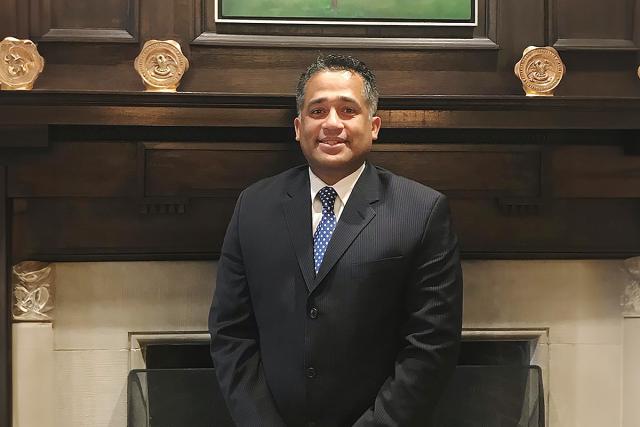 Alex Guzman, wearing a blue suit, stands in front of a fireplace.