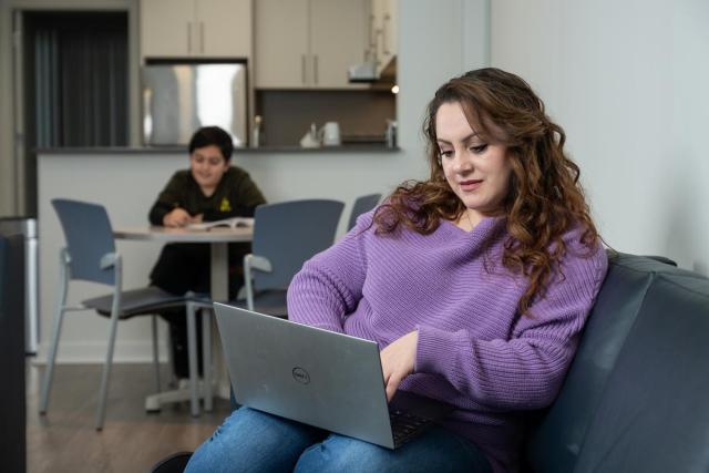 A woman works on her laptop. In the background, a young boy works at a table.