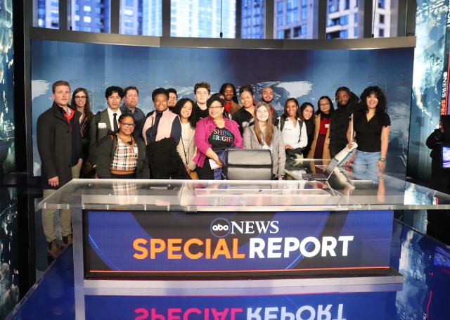 Group of students with diverse backgrounds behind a news desk standing smiling at the camera