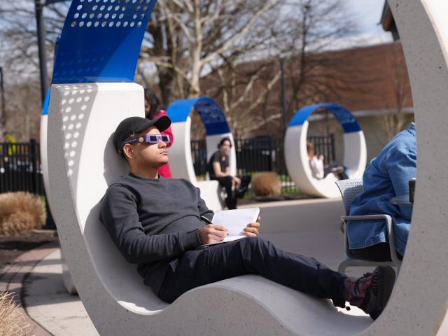 Male wearing gray shirt, holding a notebook wearing solar eclipse glasses and a black hat sitting on a chair 