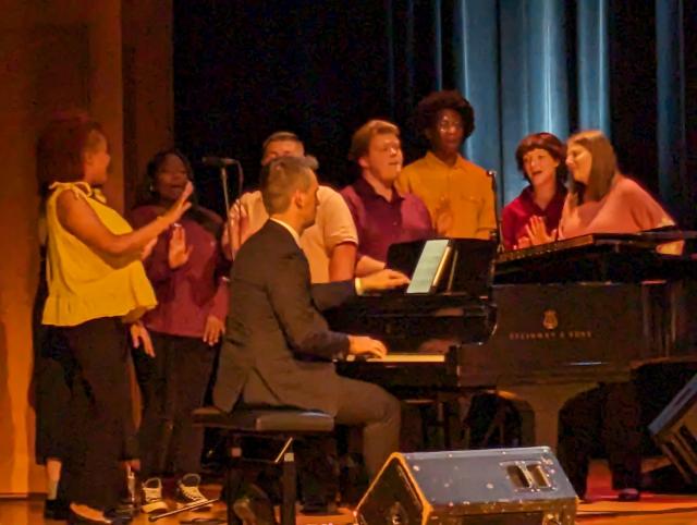 A closeup of Kean's Broadway Babies on stage, singing around a piano.