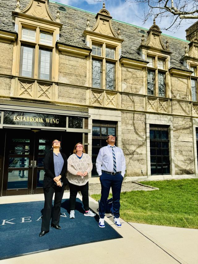 Dr. Repollet, Chief of Staff Audrey Kelly and Executive Assistant Maris Henson, wearing protective glasses, gaze up at the eclipse outside Kean Hall.