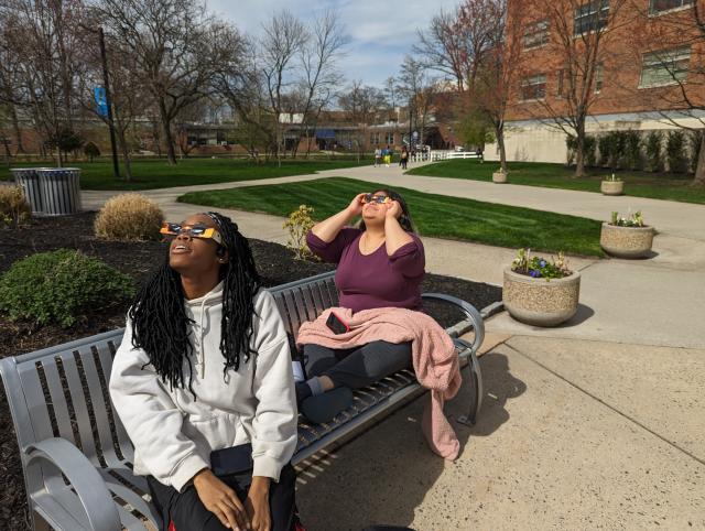 Two women, one with a gray sweater, purple shirt, with the eclipse glasses looking up