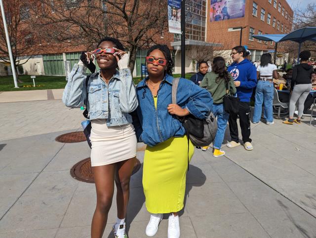Two black women wearing blue jean jeackets and one white dress and a yellow dress looking up at the solar eclipse with the glasses on
