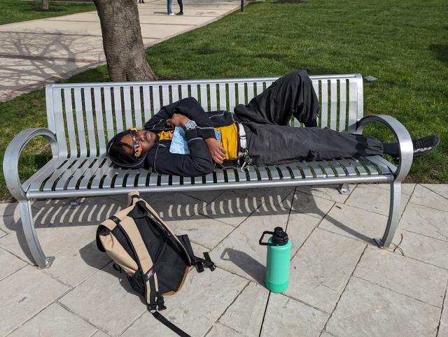 A black male laying down wearing black jeans looking up with his solar eclipse glasses crossing his arms