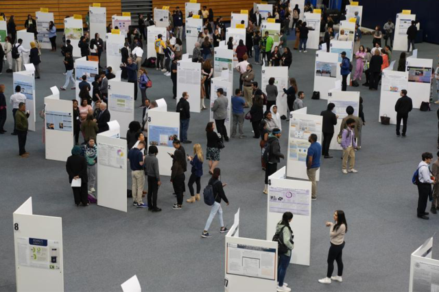 Students stand before their research poster boards, explaining their research to Research Days' visitors.