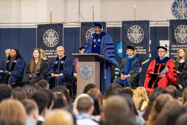 A group of diverse professionals on stage wearing colorful gowns speaking to a group of students 