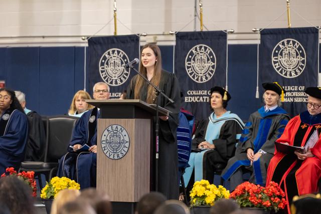 A speaker with brown hair wearing a black gown speaking on stage to students