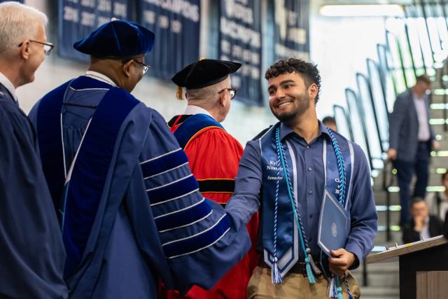 A male student wearing a stole with brown curly hair shaking hands with Kean President wearing a blue gown and 