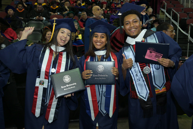 Three Kean University graduates hold their diploma covers and smile at Commencement.