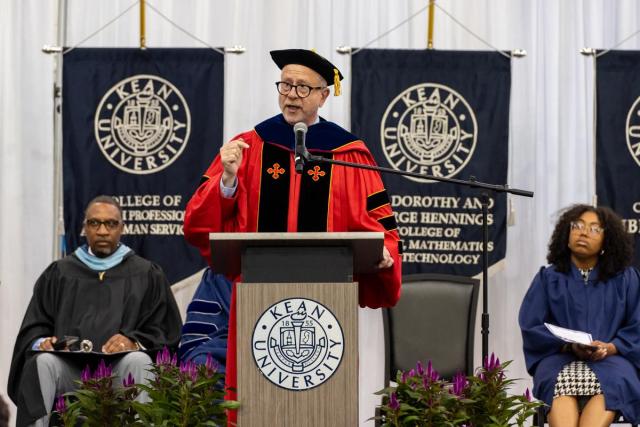 Provost David Birdsell, in a red commencement gown, speaks behind a podium.