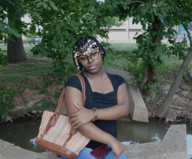 Photo of Faith Taylor in front of a creek. Faith's arms are crossed while holding her bag as she smiles at the camera.