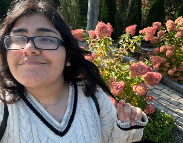 Photo of Ashley Acevedo in front of peach-colored flowers. Ashley wears glasses and a sweater as they smile at the camera.