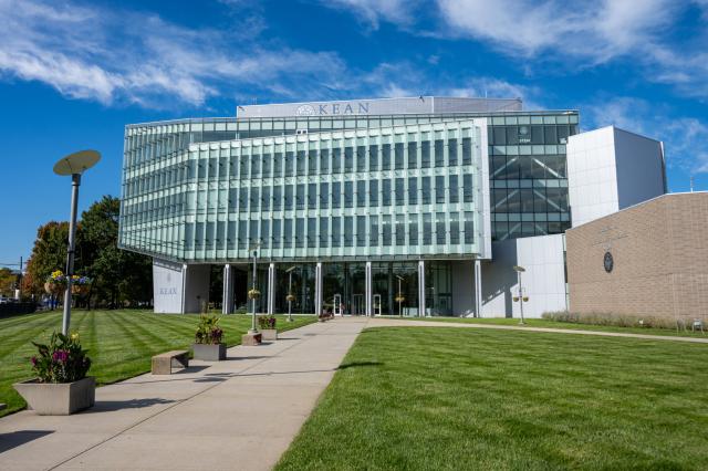 Outside of a large, six-story building lined with light green windows; green grass and blue skies.