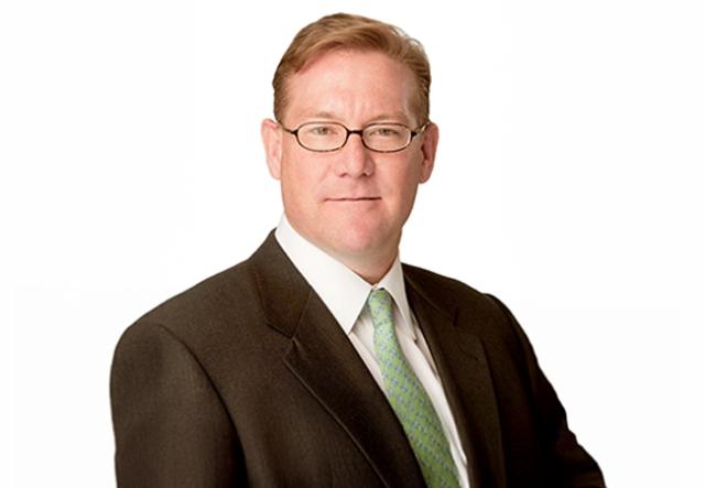 Headshot of a white male with light brown-reddish hair, glasses, wearing a dark suit, white shirt and light green tie.