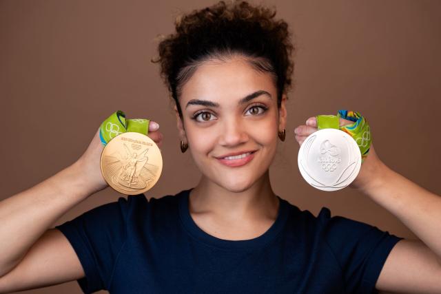 Smiling young Hispanic female holding two Olympic medals on either side of her face; she has short curly hair and is wearing a blue shirt.