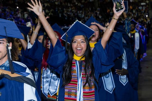 Excited female graduate in blue cap and gown, colorful stole, arms in the air. 