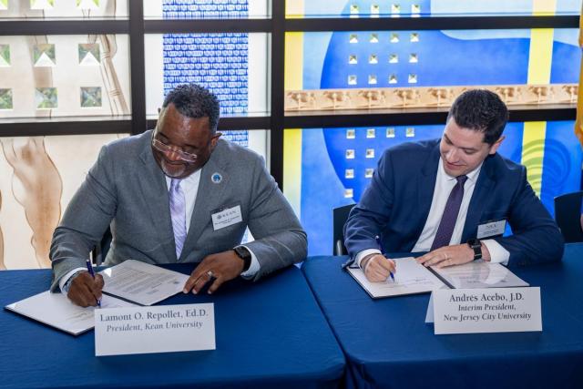 Black male wearing a gray suit and Hispanic male wearing a blue suit, side by side, each signing a formal document at a blue table; behind them is a stained glass window.