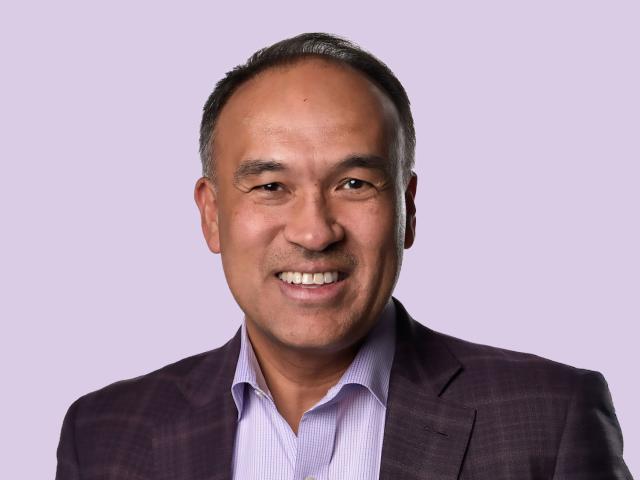 Mark Tatum smiling in a dark blazer and light purple shirt against an orange-to-green gradient background.