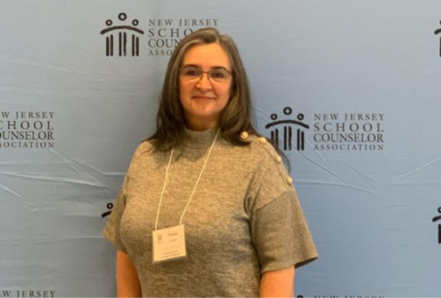 Woman with brown hair, glasses, a tan shirt and a conference name badge, in front of a light blue background
